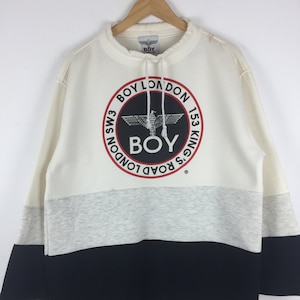 Könnte beinhalten: Ein farbblockierter Pullover in Creme, Grau und Schwarz. Der Pullover hat eine große kreisförmige Grafik mit dem Text "BOY LONDON" und einem Adleremblem. Das Kleidungsstück hängt an einem Holzbügel.
