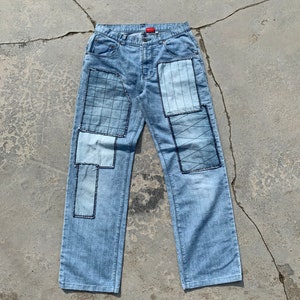 Puede incluir: Vaqueros de mezclilla azul claro con múltiples paneles de patchwork cosidos en la parte delantera de las piernas. Los paneles están hechos de mezclilla azul de diferentes tonos y tienen costuras negras.