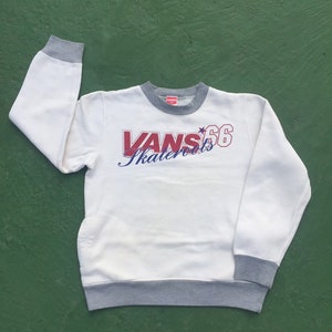 May include: White crew neck sweatshirt with gray trim on the collar, cuffs, and hem. The front features the text "VANS Skateboards 66" in red and blue script and block lettering. The sweatshirt is made of a soft material.