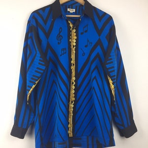 May include: A long-sleeved button-down shirt with a vibrant blue and black geometric pattern. The shirt features musical notes and a gold-colored flute-like design down the front. The collar and cuffs are black, and the shirt is hanging on a wooden hanger.