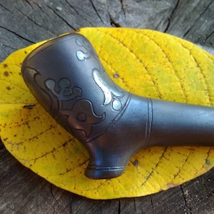 Op de afbeelding: Een zwarte keramische pijp met een zilveren bloemenmotief. De pijp ligt op een geel blad.