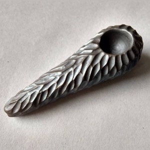 May include: A gray ceramic pipe with a textured, leaf-like design. The pipe has a round bowl and a long, narrow stem.