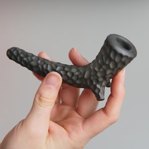 Puede incluir: Una pipa de cerámica negra con una superficie texturizada. La pipa tiene forma de rama con una cazoleta redonda en el extremo.