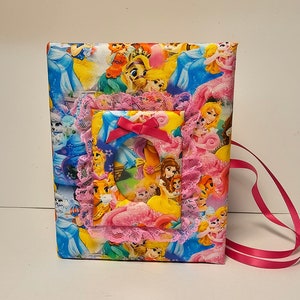 Puede incluir: Un &aacute;lbum de fotos de tela rosa y azul con un tema de princesas. El &aacute;lbum tiene un marco incorporado con una imagen de princesas Disney. El &aacute;lbum tiene una cinta rosa.