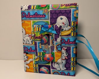 May include: A colorful fabric-covered journal with a Dalmatian dog theme. The cover features a variety of Dalmatian puppies, bones, and balls. The journal has a blue ribbon closure.