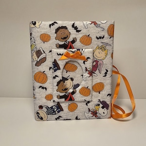 Puede incluir: Un &aacute;lbum de fotos de tela gris con tem&aacute;tica de Halloween con personajes de Peanuts. El &aacute;lbum tiene una ventana recortada y una cinta naranja.