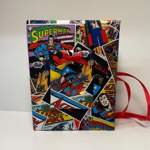 May include: A fabric-covered box featuring a collage of Superman comic book panels. The box is adorned with images of Superman in action, with vibrant colors and the iconic "Superman" logo. A red ribbon is tied around the box.