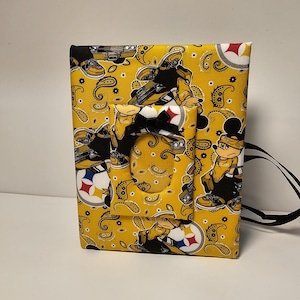 May include: A yellow fabric pouch with a black strap. The pouch has a paisley pattern with cartoon characters wearing Pittsburgh Steelers gear. The pouch has a clear plastic window.
