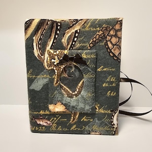 Puede incluir: Un diario de tela gris oscuro con cierre de cinta marrón. La portada presenta un estampado vintage de criaturas marinas, incluyendo un pulpo, una estrella de mar y tortugas marinas. El estampado es en tonos dorados, marrones y blancos.