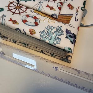 Puede incluir: Un &aacute;lbum de recortes de tela blanca con un tema n&aacute;utico. La portada presenta una variedad de ilustraciones de vida marina, incluyendo peces, anclas y un salvavidas. El &aacute;lbum de recortes est&aacute; encuadernado con una cinta azul.