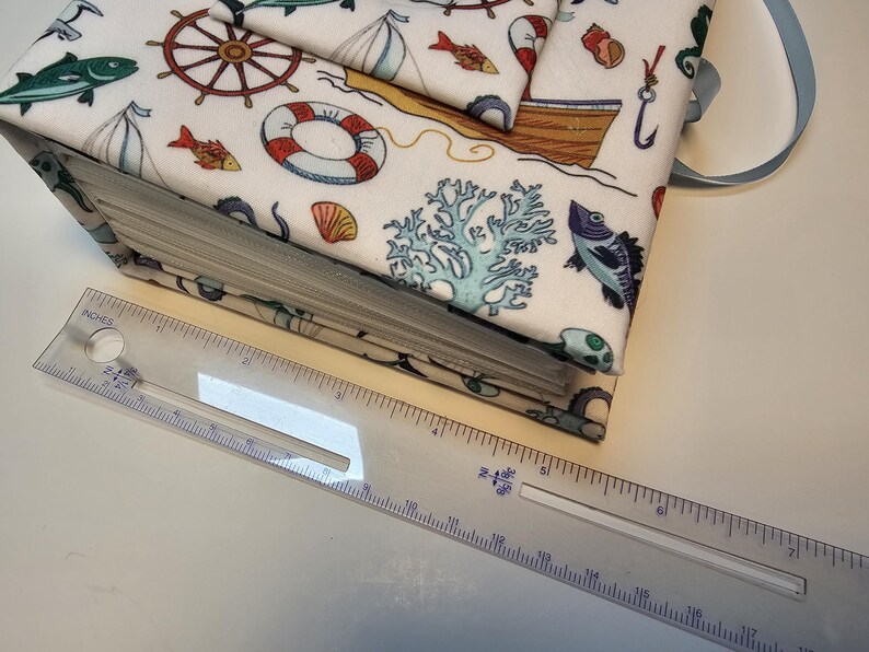 Puede incluir: Un &aacute;lbum de recortes de tela blanca con un tema n&aacute;utico. La portada presenta una variedad de ilustraciones de vida marina, incluyendo peces, anclas y un salvavidas. El &aacute;lbum de recortes est&aacute; encuadernado con una cinta azul.