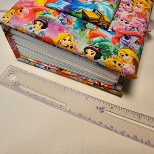 Puede incluir: Un &aacute;lbum de fotos cubierto de tela colorida con un tema de princesas. El &aacute;lbum est&aacute; cerrado y presenta im&aacute;genes de princesas de Disney como Blancanieves, Cenicienta y Rapunzel. Una regla se coloca debajo del &aacute;lbum para mostrar su tama&ntilde;o.