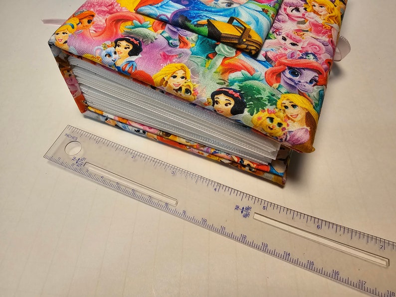 Puede incluir: Un &aacute;lbum de fotos cubierto de tela colorida con un tema de princesas. El &aacute;lbum est&aacute; cerrado y presenta im&aacute;genes de princesas de Disney como Blancanieves, Cenicienta y Rapunzel. Una regla se coloca debajo del &aacute;lbum para mostrar su tama&ntilde;o.