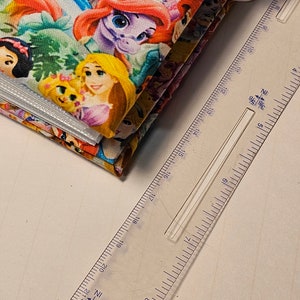 Puede incluir: Una pila de cuadrados de tela coloridos con im&aacute;genes de princesas Disney, incluyendo Ariel, Bella, Blancanieves y Rapunzel. La tela est&aacute; doblada y una regla se coloca al lado.