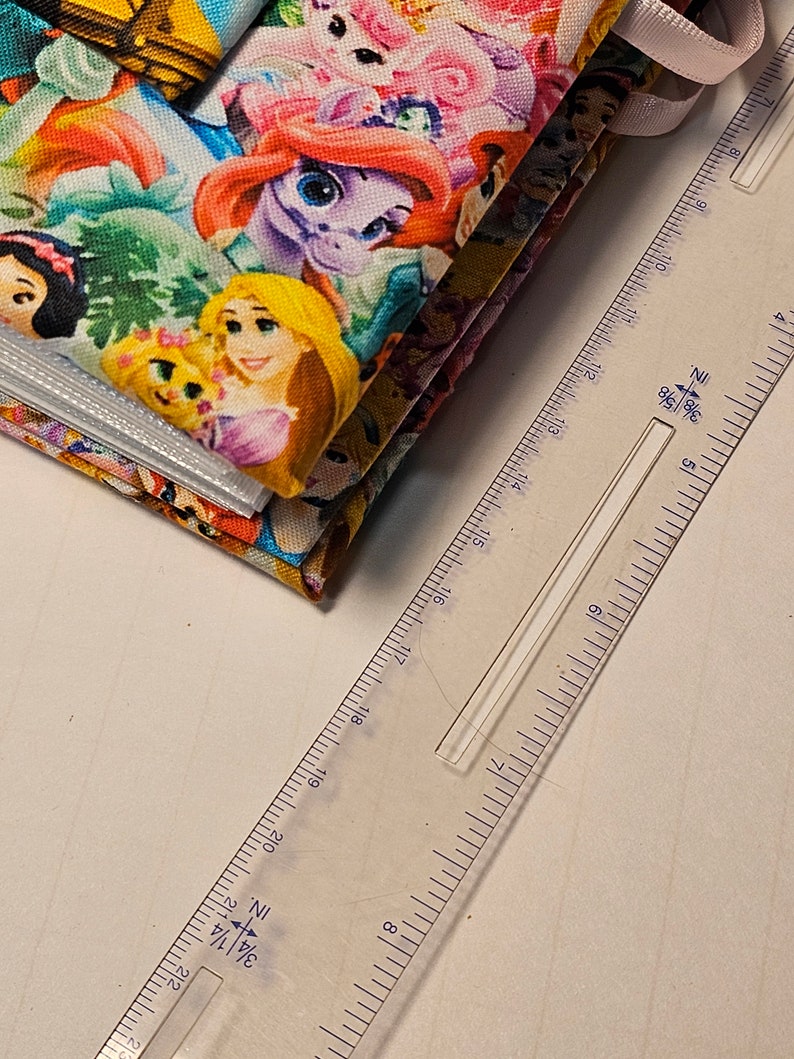 Puede incluir: Una pila de cuadrados de tela coloridos con im&aacute;genes de princesas Disney, incluyendo Ariel, Bella, Blancanieves y Rapunzel. La tela est&aacute; doblada y una regla se coloca al lado.