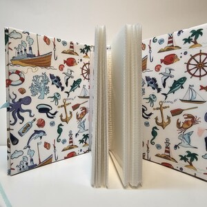 Puede incluir: Un &aacute;lbum de fotos blanco con tem&aacute;tica n&aacute;utica. La portada presenta una variedad de ilustraciones, incluyendo un barco, anclas, peces y conchas marinas. El &aacute;lbum es perfecto para guardar recuerdos de unas vacaciones en la playa.