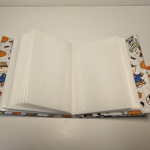 Puede incluir: Un &aacute;lbum de fotos blanco con una cubierta de tela gris con un tema de Halloween de Peanuts. La cubierta tiene im&aacute;genes de Snoopy, Woodstock y otros personajes de Peanuts vestidos con disfraces de Halloween. El &aacute;lbum tiene un cierre de cinta.