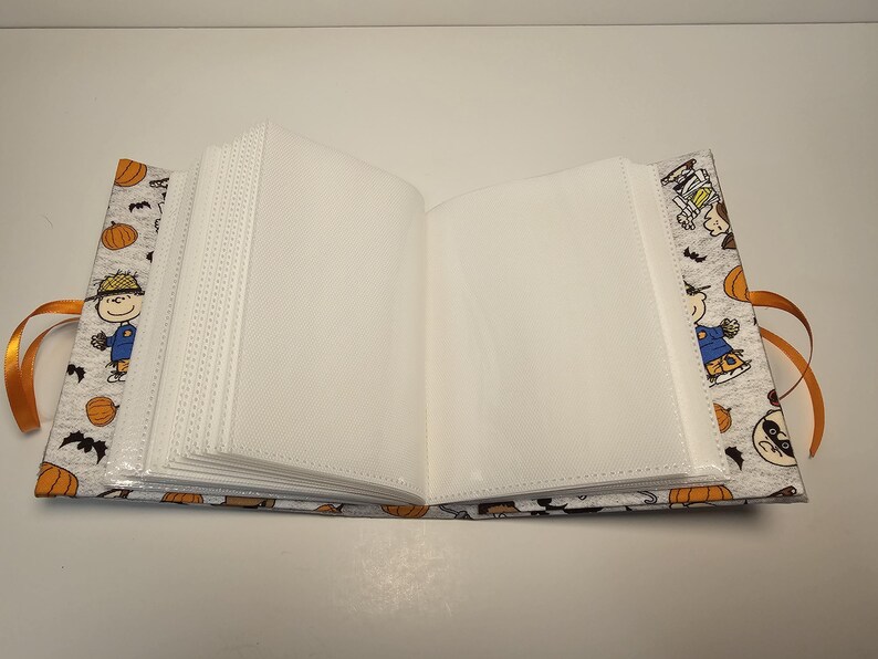 Puede incluir: Un &aacute;lbum de fotos blanco con una cubierta de tela gris con un tema de Halloween de Peanuts. La cubierta tiene im&aacute;genes de Snoopy, Woodstock y otros personajes de Peanuts vestidos con disfraces de Halloween. El &aacute;lbum tiene un cierre de cinta.