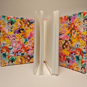 Puede incluir: Un &aacute;lbum de fotos rosa con una cubierta de tela colorida con princesas Disney y ponis. El &aacute;lbum tiene un cierre de cinta rosa.