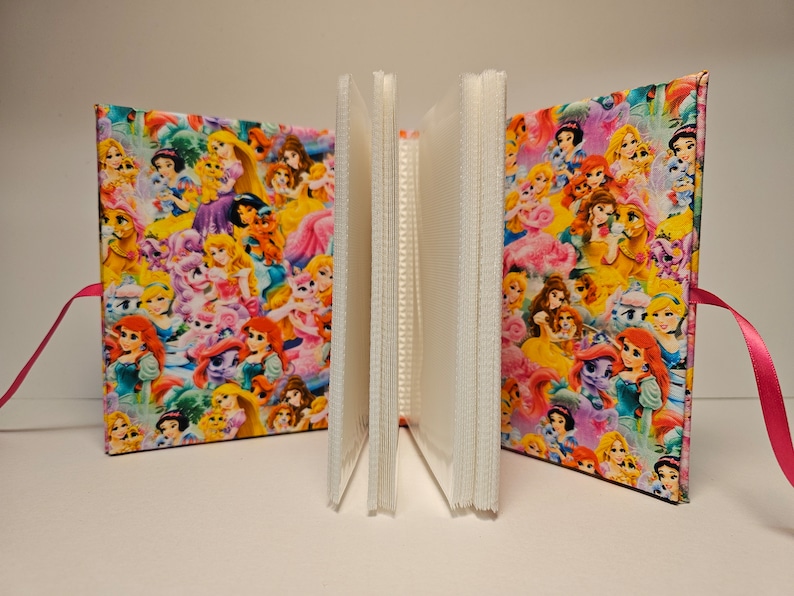 Puede incluir: Un &aacute;lbum de fotos rosa con una cubierta de tela colorida con princesas Disney y ponis. El &aacute;lbum tiene un cierre de cinta rosa.