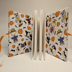 Puede incluir: Un &aacute;lbum de recortes de tela blanca con lazos de cinta naranja. La portada presenta un patr&oacute;n repetitivo de personajes de Peanuts vestidos con disfraces de Halloween, calabazas y murci&eacute;lagos.