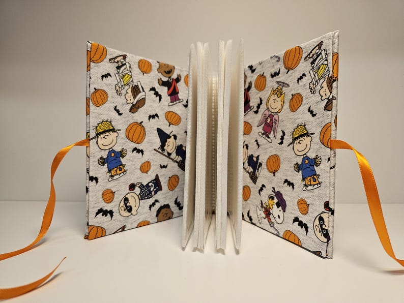 Puede incluir: Un &aacute;lbum de recortes de tela blanca con lazos de cinta naranja. La portada presenta un patr&oacute;n repetitivo de personajes de Peanuts vestidos con disfraces de Halloween, calabazas y murci&eacute;lagos.