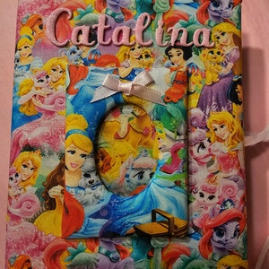 Puede incluir: Un &aacute;lbum de fotos de tela rosa con tem&aacute;tica de princesas y unicornios. El &aacute;lbum tiene un recorte para una foto y el nombre "Catalina" est&aacute; bordado en la parte delantera.