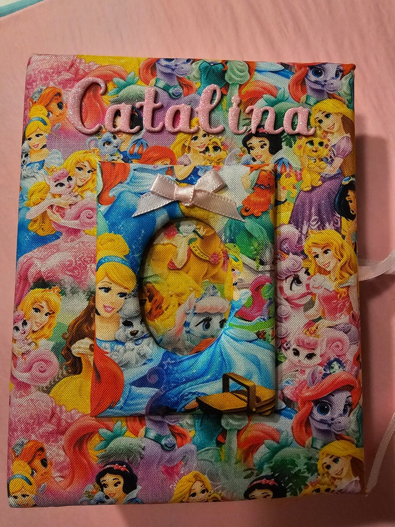 Puede incluir: Un &aacute;lbum de fotos de tela rosa con tem&aacute;tica de princesas y unicornios. El &aacute;lbum tiene un recorte para una foto y el nombre "Catalina" est&aacute; bordado en la parte delantera.