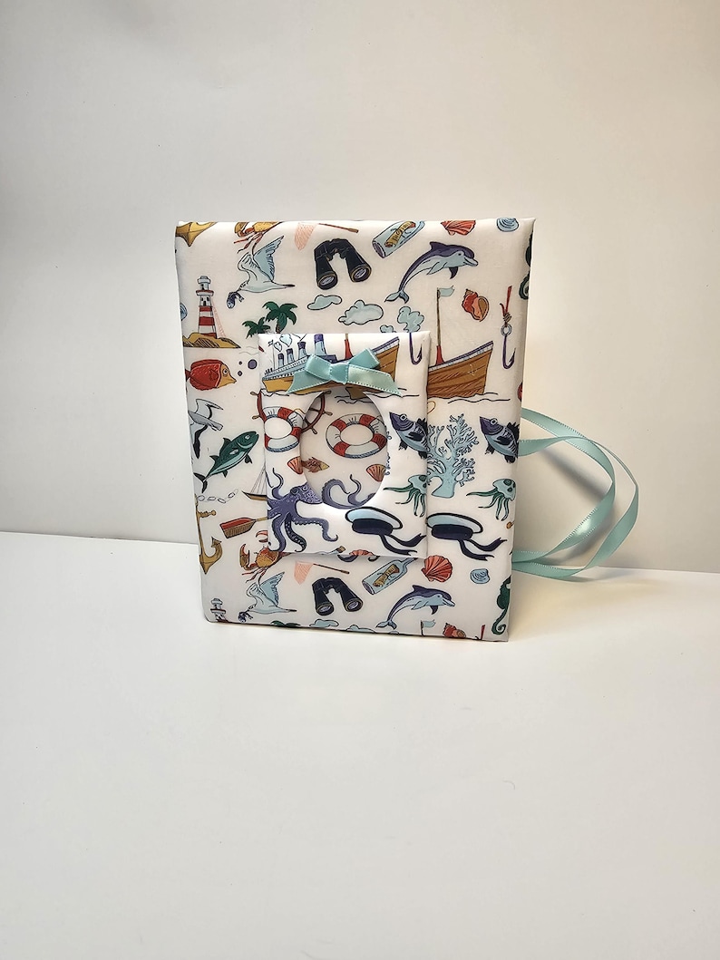 Puede incluir: Un &aacute;lbum de fotos blanco con una cinta azul. La portada presenta un tema n&aacute;utico con ilustraciones de criaturas marinas, barcos y otros objetos n&aacute;uticos.