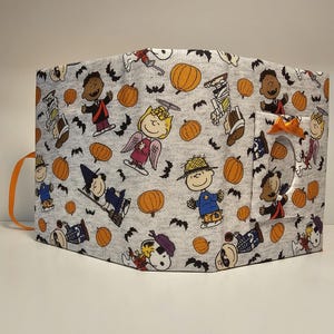 Puede incluir: Una cubierta de libro de tela gris con tem&aacute;tica de Halloween. La cubierta presenta personajes de Peanuts vestidos con disfraces, calabazas y murci&eacute;lagos. La cubierta tiene un bolsillo en el lateral con un lazo naranja.