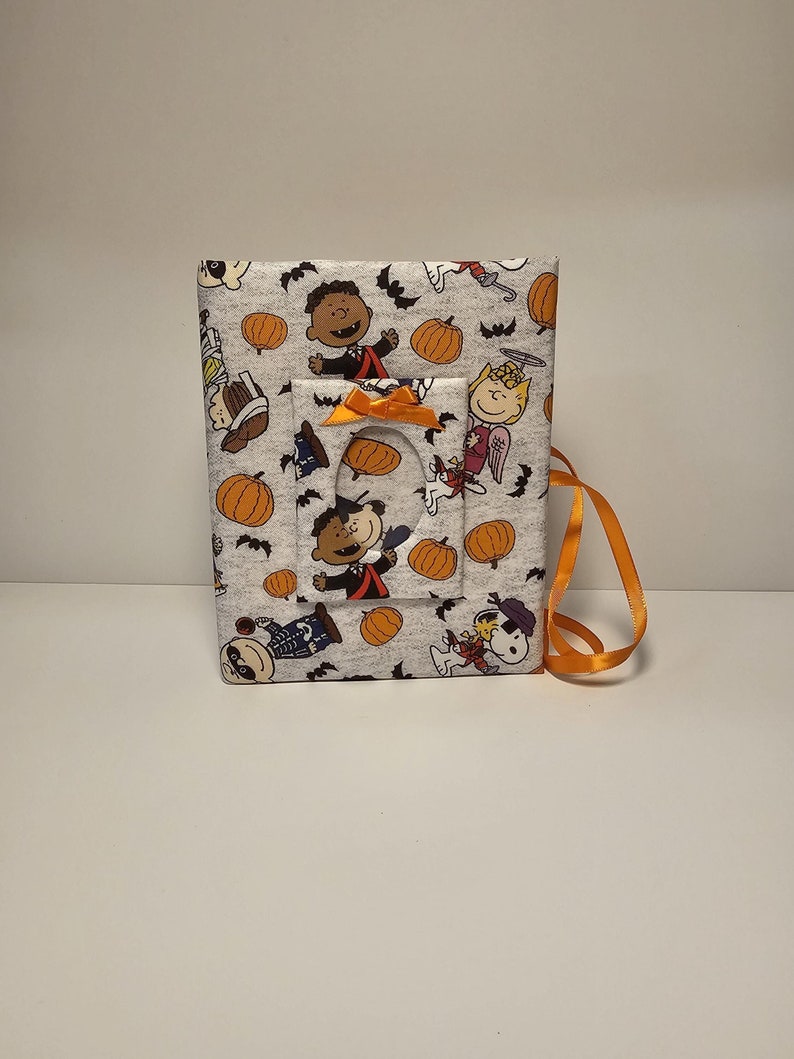 Puede incluir: Un &aacute;lbum de fotos de tela gris con tem&aacute;tica de Halloween con personajes de Peanuts. El &aacute;lbum tiene una ventana recortada y una cinta naranja.
