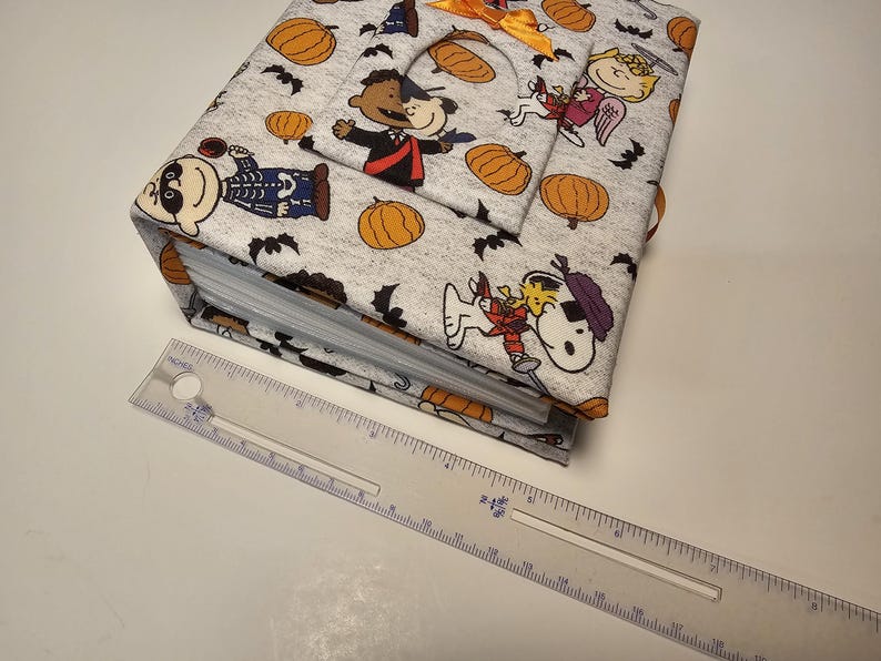 Puede incluir: Un &aacute;lbum de fotos de tela gris con tem&aacute;tica de Halloween. La portada presenta personajes de Peanuts disfrazados y calabazas. El &aacute;lbum tiene una encuadernaci&oacute;n de anillas y est&aacute; cerrado.