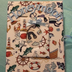 Puede incluir: Un &aacute;lbum de fotos de tela blanca con tem&aacute;tica n&aacute;utica. El &aacute;lbum tiene una cinta azul y un peque&ntilde;o marco con una ventana transparente. El &aacute;lbum tiene la palabra "honeymoon" escrita en la parte delantera en letras de purpurina azul.