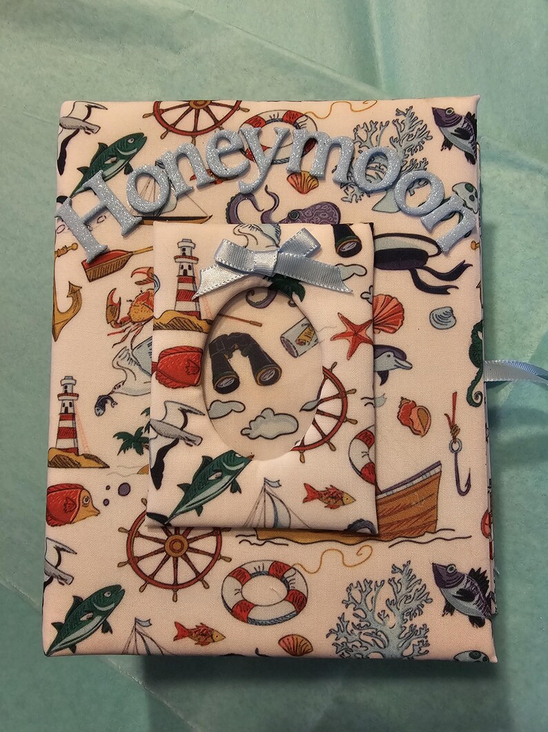 Puede incluir: Un &aacute;lbum de fotos de tela blanca con tem&aacute;tica n&aacute;utica. El &aacute;lbum tiene una cinta azul y un peque&ntilde;o marco con una ventana transparente. El &aacute;lbum tiene la palabra "honeymoon" escrita en la parte delantera en letras de purpurina azul.