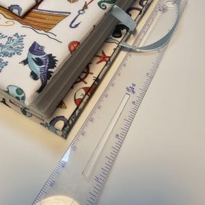 Puede incluir: Un diario blanco cubierto de tela con un marcap&aacute;ginas de cinta azul. La portada presenta un tema n&aacute;utico con ilustraciones de peces, conchas, anclas y un barco. El diario se mide con una regla, mostrando que mide aproximadamente 20 cm de largo.