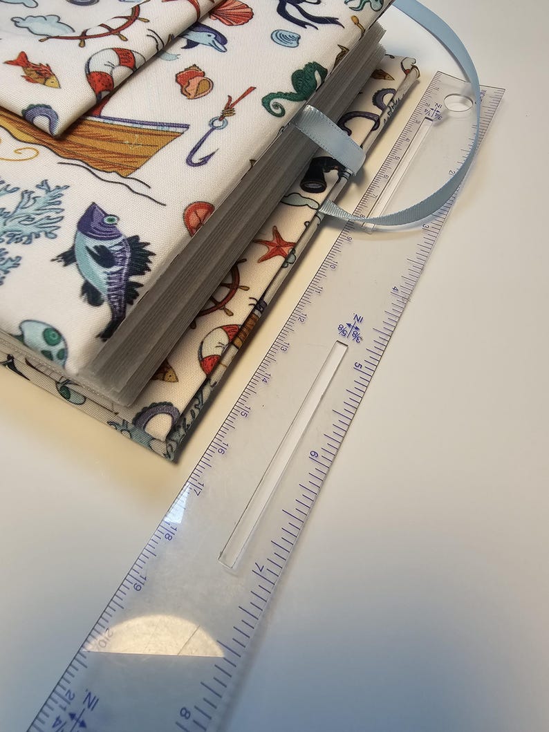 Puede incluir: Un diario blanco cubierto de tela con un marcap&aacute;ginas de cinta azul. La portada presenta un tema n&aacute;utico con ilustraciones de peces, conchas, anclas y un barco. El diario se mide con una regla, mostrando que mide aproximadamente 20 cm de largo.