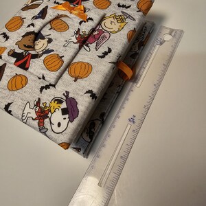 Puede incluir: Un diario de tela gris y blanco con tem&aacute;tica de Halloween. La portada presenta personajes de Peanuts disfrazados, calabazas y murci&eacute;lagos. El diario est&aacute; cerrado y tiene una cinta naranja.