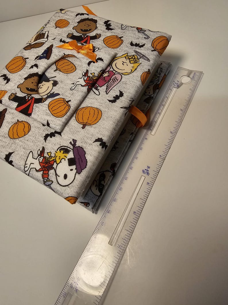 Puede incluir: Un diario de tela gris y blanco con tem&aacute;tica de Halloween. La portada presenta personajes de Peanuts disfrazados, calabazas y murci&eacute;lagos. El diario est&aacute; cerrado y tiene una cinta naranja.