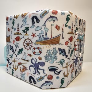 Puede incluir: Un contenedor de almacenamiento de tela blanca con un tema n&aacute;utico. El contenedor est&aacute; decorado con una variedad de criaturas marinas, barcos y otras im&aacute;genes n&aacute;uticas. El contenedor tiene un asa a rayas azul y blanca.