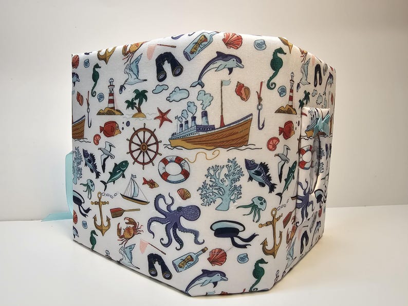 Puede incluir: Un contenedor de almacenamiento de tela blanca con un tema n&aacute;utico. El contenedor est&aacute; decorado con una variedad de criaturas marinas, barcos y otras im&aacute;genes n&aacute;uticas. El contenedor tiene un asa a rayas azul y blanca.