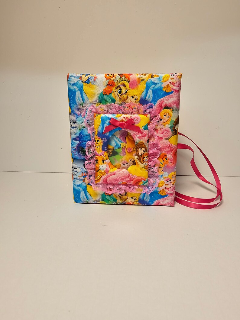 Puede incluir: Un &aacute;lbum de fotos de tela rosa y azul con un tema de princesas. El &aacute;lbum tiene un marco incorporado con una imagen de princesas Disney. El &aacute;lbum tiene una cinta rosa.