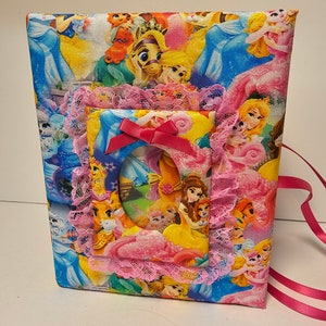 Puede incluir: Un &aacute;lbum de fotos de tela rosa y blanco con un tema de princesa. El &aacute;lbum tiene un borde de encaje y un lazo rosa. La portada presenta una variedad de princesas y personajes de Disney.
