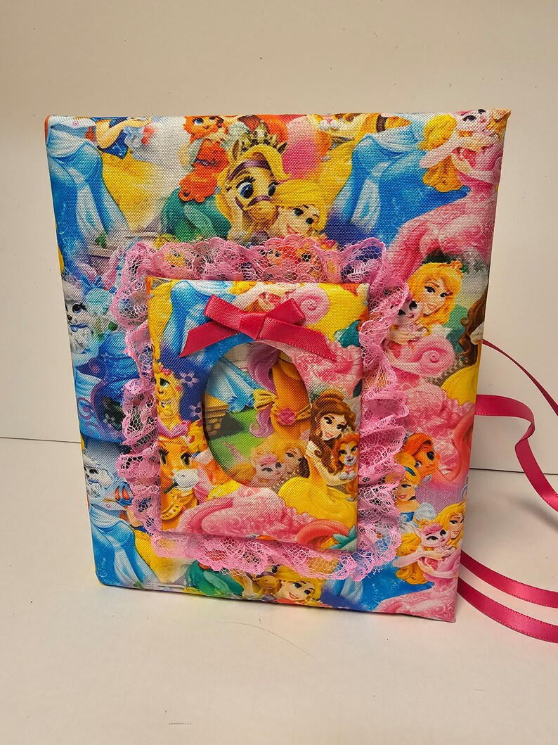 Puede incluir: Un &aacute;lbum de fotos de tela rosa y blanco con un tema de princesa. El &aacute;lbum tiene un borde de encaje y un lazo rosa. La portada presenta una variedad de princesas y personajes de Disney.
