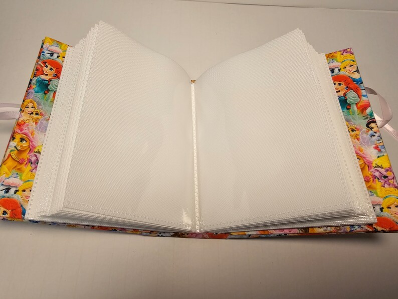 Puede incluir: Un &aacute;lbum de fotos blanco con fundas de pl&aacute;stico transparente para guardar fotos. La cubierta est&aacute; hecha de tela y presenta una impresi&oacute;n colorida de princesas Disney.
