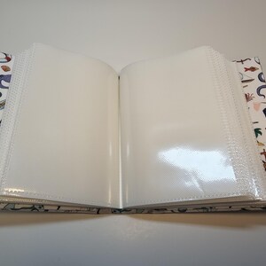 Puede incluir: Un &aacute;lbum de fotos blanco con tem&aacute;tica n&aacute;utica. La portada presenta ilustraciones de un pulpo, un barco, un salvavidas y otros objetos n&aacute;uticos. El &aacute;lbum est&aacute; abierto para revelar p&aacute;ginas de pl&aacute;stico transparente para guardar fotos.