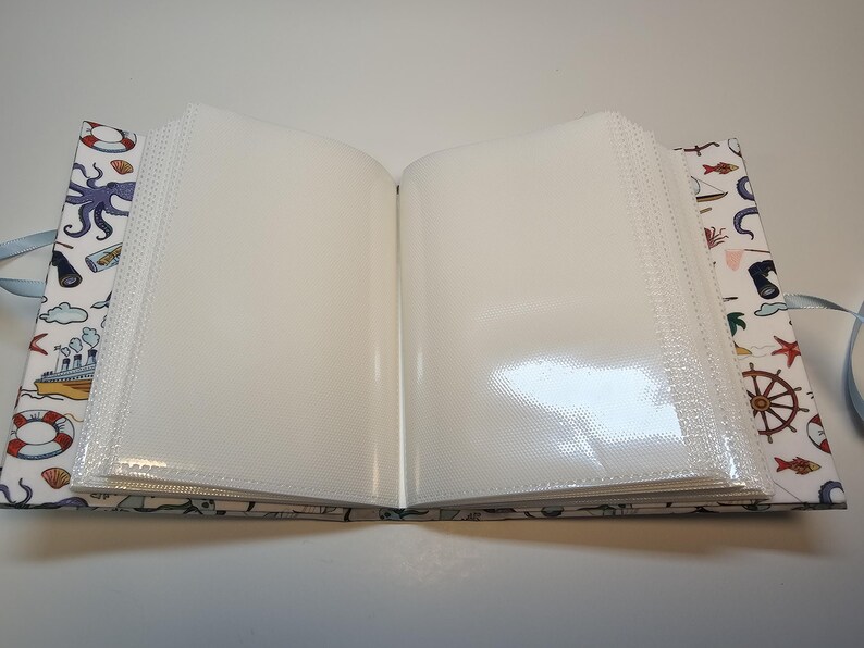 Puede incluir: Un &aacute;lbum de fotos blanco con tem&aacute;tica n&aacute;utica. La portada presenta ilustraciones de un pulpo, un barco, un salvavidas y otros objetos n&aacute;uticos. El &aacute;lbum est&aacute; abierto para revelar p&aacute;ginas de pl&aacute;stico transparente para guardar fotos.