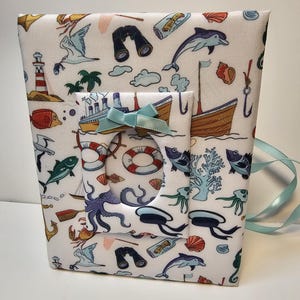 Puede incluir: Un &aacute;lbum de fotos de tela blanca con tem&aacute;tica n&aacute;utica. La portada presenta una variedad de ilustraciones con tem&aacute;tica mar&iacute;tima, incluyendo un barco, un pulpo, un faro y un delf&iacute;n. El &aacute;lbum tiene una cinta azul.