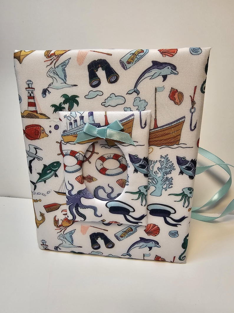 Puede incluir: Un &aacute;lbum de fotos de tela blanca con tem&aacute;tica n&aacute;utica. La portada presenta una variedad de ilustraciones con tem&aacute;tica mar&iacute;tima, incluyendo un barco, un pulpo, un faro y un delf&iacute;n. El &aacute;lbum tiene una cinta azul.