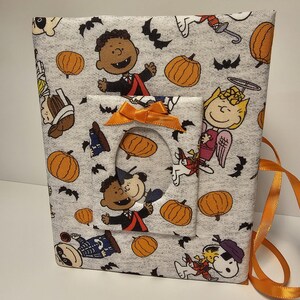 Puede incluir: Un marco de fotos de tela gris con tem&aacute;tica de Halloween. El marco presenta un patr&oacute;n de personajes de Peanuts con calabazas, murci&eacute;lagos y personajes vestidos con disfraces. El marco tiene un recorte para una foto y una cinta naranja brillante.