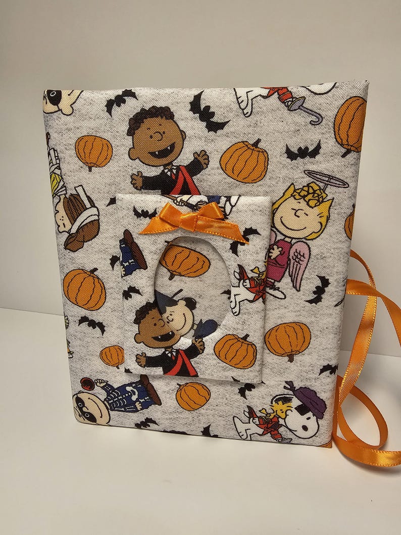 Puede incluir: Un marco de fotos de tela gris con tem&aacute;tica de Halloween. El marco presenta un patr&oacute;n de personajes de Peanuts con calabazas, murci&eacute;lagos y personajes vestidos con disfraces. El marco tiene un recorte para una foto y una cinta naranja brillante.
